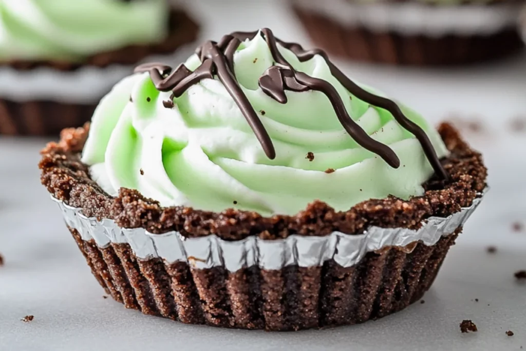 Close-up of a mini grasshopper pie with a chocolate cookie crust, light green mint cream filling swirled on top, and a drizzle of dark chocolate, served in a foil tart wrapper on a clean surface.