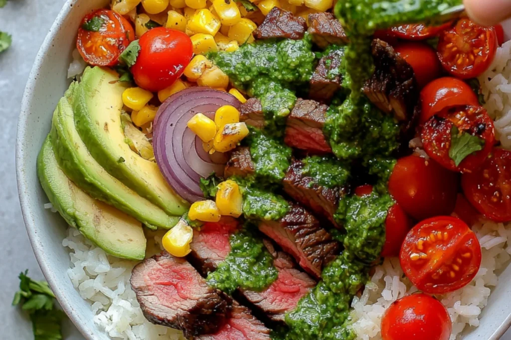Close-up of a steak rice bowl topped with avocado slices, cherry tomatoes, roasted corn, red onion, and green cilantro cream sauce being drizzled over sliced medium-rare steak.